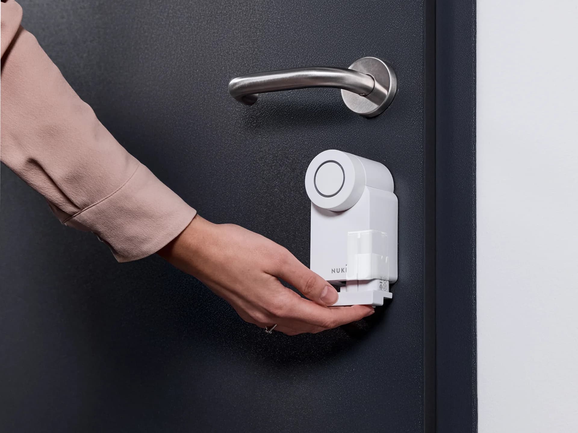Bir kadın, gümüş kollu koyu renk bir kapıdaki beyaz Nuki Smart Lock kilidine Power Pack'i yerleştiriyor